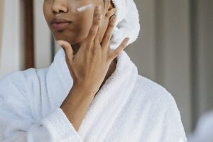Crop anonymous female in white bathrobe with towel on head applying facial moisturizing cream on face while standing in bathroom